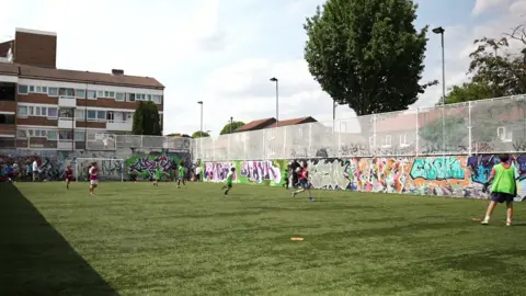 Children play football on the pitch, which has graffiti on the walls