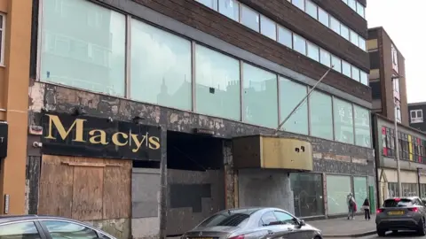BBC The front of a former hotel building, looking worse for wear. It has boarded up fronts and a row of large glass windows. Cars are parked on the road in front.