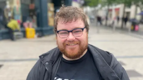 Paul Maye is wearing a black coat and black t-shirt, he has black glasses on. He has ginger facial hair and brown head hair. The background is blurry.