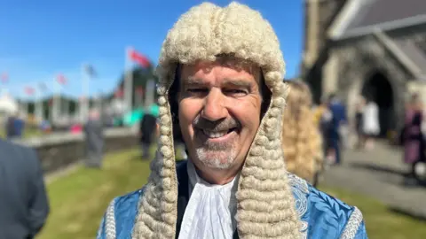 Laurence, un hombre con barba gris, viste una larga peluca ceremonial blanca y una túnica azul, y hay una iglesia y una hilera de banderas al fondo.