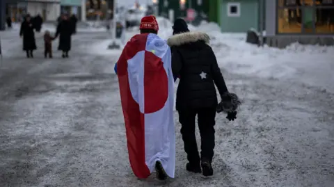 Two people walk down an icy street. One is wearing a draped Greenland flag.