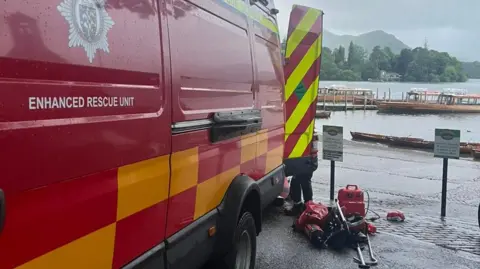 A red emergency rescue unit is parked beside a pier at Derwent Water. There are other boats tied up to the pier with the Lake District fells in the background.
