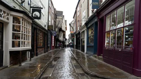 A picture of the Shambes in York, a historic street. The street is narrow and cobbled with shops lining either side. 