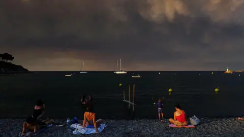 Getty Images O fundo da imagem mostra várias pessoas sentadas na praia com o mar à sua frente com alguns barcos e o céu cheio de fumaça preta grossa