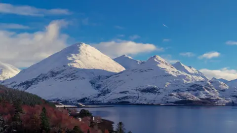 John Pullen Two snow-capped mountains behind a body of water. There is a collection of trees in the foreground of the image which are red and brown. A still body of water is in the centre and towards the back of the image. The sky above is blue with puffy white clouds.