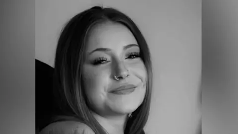 A head and shoulders black and white image of a young woman who is smiling. She has a nose ring and is looking into the camera.