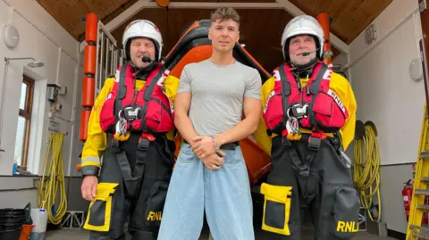 Ren Gill stood in between two RNLI crew members in full water rescue equipement at Beaumaris station on Anglesey in June 2023