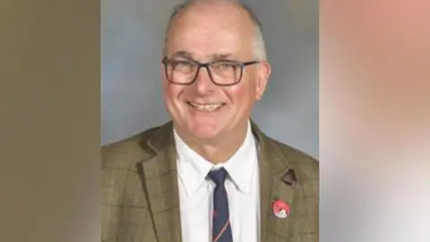 An older man in glasses, smiling. He's wearing a tweed jacket and a shirt and tie.