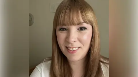 Nicola Key A woman with long brown hair and a white shirt smiles as she takes a selfie while standing inside a house.