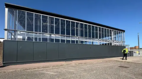 Reprezent Project A hoarding panel along the bottom of a railway station building is painted a dark grey colour and a man wearing a high-visibility jacket walks past the structure.