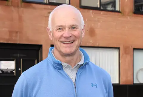 DAGCAS A man with a blue jumper, standing outside a sandstone building. He has grey hair and is smiling broadly. 