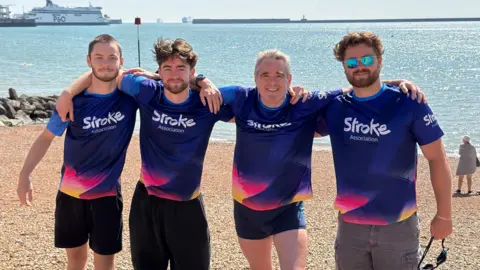 Four men in purple Stroke Association tops stand on a beach.