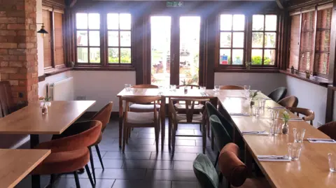 Bev Rimmer/BBC Sunlight streams in through a set of French doors and windows, onto a pub dining room that has four tables set out around the dining space. Each wooden, pine-coloured table has drinking glasses, napkins, and cutlery laid out, and a chair at each place. The left-most table has a small pot of pink flowers in the middle. 