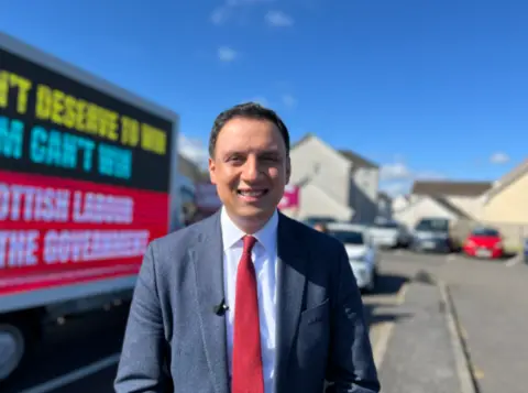 Man in a suit and red tie smiling at the camera on a residential street, with a campaign billboard truck behind him displaying political messaging in bright colours