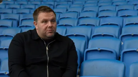 Russ Wood, who has grey/brown hair and stubble and wears a black, zip up jacket, sits on a blue stadium chair with rows of empty blue chairs behind him.