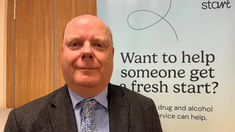 Craig Blundred, Director of Public Health, Hartlepool Borough Council, standing in front of a drug advice poster, slightly smiling, wearing a suit and tie.