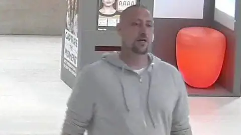 Thames Valley Police A man is walking through a shopping centre in a grey hooded top. He has short hair and a short goatee beard.