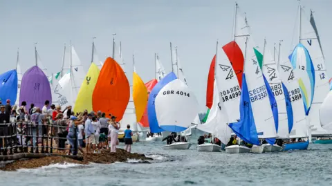 A line of small yachts with multi-coloured sails sailing close to the shore where crowds of spectators are watching