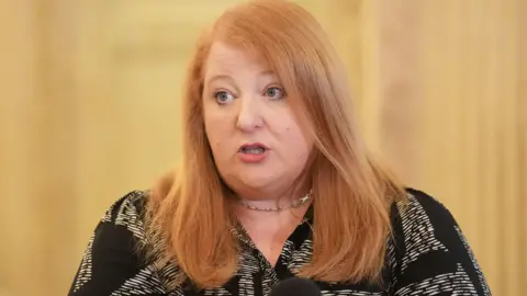 PA Media Naomi Long with long red hair. She is wearing a black blouse with a cream pattern. She is wearing a necklace.