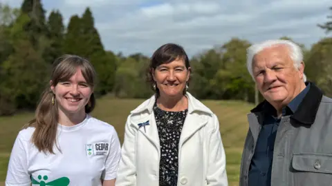 Amy Laird is on the left, Helen James is in the middle and Roy Anderson on the right. Amy is wearing a white t-shirt with a green insect and black CEDaR logo. Helen is wearing a black top with flowers and a white jacket with an insect brooch. Roy is wearing a grey jacket with a navy shirt. Green trees and grass are in the background. 