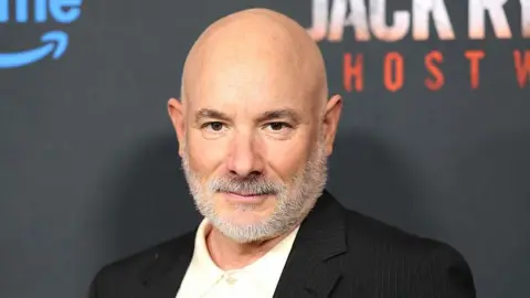 Getty Images A bald man poses for photos during a film premiere. He has a thick grey beard and wears a black suit jacket with a white top underneath. 