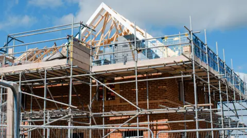 A stock image of a house being built with scaffolding around the roof and beams in place on top of red brick walls.