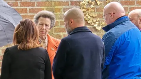 Princess Anne is wearing an orange coat and is talking to four people in a garden in Speke