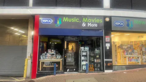 BBC The RSPCA's Music, Movies and More shop which has its lights switched off. There is a large loading bay with shutters to the left hand side and another charity shop to the right hand side. 