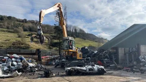 Gemma Dillon/BBC A crane towers over a crushed vehicle at a scrapyard. In the background are green fields and trees.