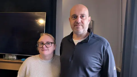 Charlotte and Carl Cooper. Charlotte is wearing a beige knitted jumper, Carl is wearing a grey jumper over a white t-shirt. There is a large television on the wall behind them and blue curtains to the right of them.