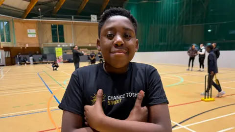 BBC/Jim Scott Shirel, who is one of the members, stands with his hands tucked under his arms while wearing a 'Chance to Shine/Street' shirt. He is standing on the court while his friends take part in a game of cricket.