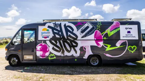 The image shows a black van with vibrant graffiti art in white, purple and lime green. The van is parked on a gravel surface. The picture was taken on a sunny day with a bright blue sky with clouds.