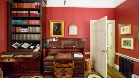 Newangle Copyright, Charles Dickens Museum A study, with red walls and dark brown furniture, including a desk and book case