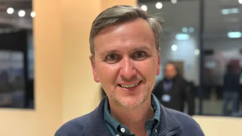 Shariqua Ahmed/BBC Andrew Pakes wearing a blue shirt. He is smiling for the camera.