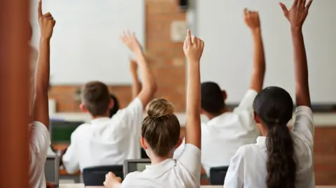 A group of students put their hands up in a room with a white board and red brick walls.