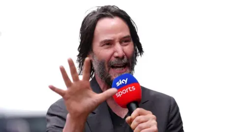 Getty Images Keanu Reeves in a black top and grey jacket. He is talking in a blue and red Sky Sports microphone and holding his hand up.
