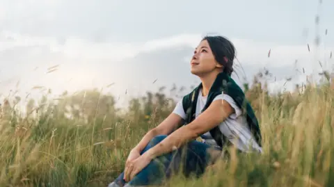 A woman is sat in a field of long grass looking at the sky. She has a white T-shirt on and a blue jumper over her shoulders with long dark hair.