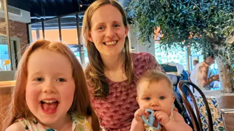 Maria with two young children sitting in a cafe. Her young daughter Emily is right up to the camera - she has auburn hair and is smiling with her mouth wide-open. A baby girl is sitting on Maria's lap, chewing on a blue toy. Maria has long brown hair and is also smiling to camera.