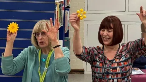 Two women sat on a chairs in a library with their arms in the air while holding what looks similar to a yellow flower in their left hand. They are telling a story to some children (not pictured). The woman on the left has blonde hair and a fringe styled into bob. She wears a turquoise top, matching love heart necklace and glasses. The woman on the right has red/brown hair styled into a bob with a fringe. She is wearing a dress that has rows of bookshelves printed onto it. 