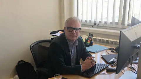 Local Democracy Reporting Service West Midlands mayor Richard Parker with his hands clasped together, wearing glasses facing the camera. He is sitting at a desk with a keyboard and paper and trays are behind him.