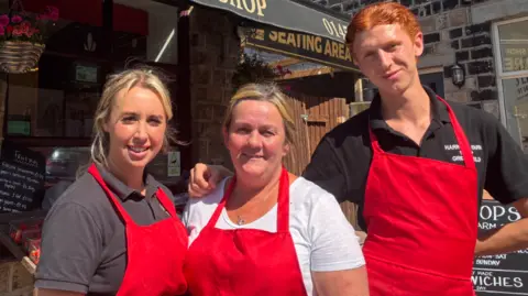 LDRS Three people wearing red aprons are standing outside a shop with black signs on a sunny day.
