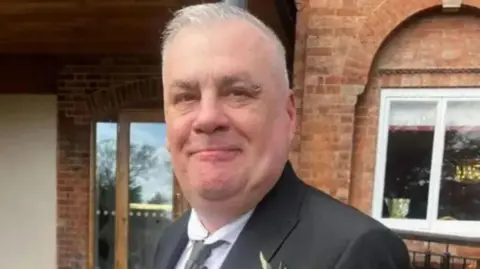 A man with short cropped grey hair and a ring piercing on the end of his left eyebrow. He is wearing a black blazer over a white shirt and a grey tie. There is a flower in the right breast pocket. He is standing in front of a red-brick building and smiling.