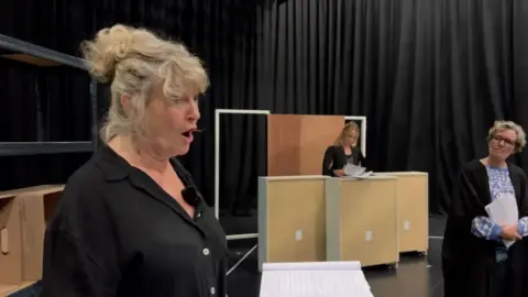 Three actors on the stage of a play about the post office scandal with black backdrop. Nearest to the camera on the left of the frame is Liz Elvin with her blonde hair tied up, she is speaking and wearing a black shirt. Behind, one actress is dressed in a blue top and black barristers robes, the third actress is behind some book shelves wearing a black top and looking at papers.