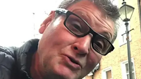 Image shows John Miller, 63, looking at the camera in black-rimmed glasses and a black jacket 