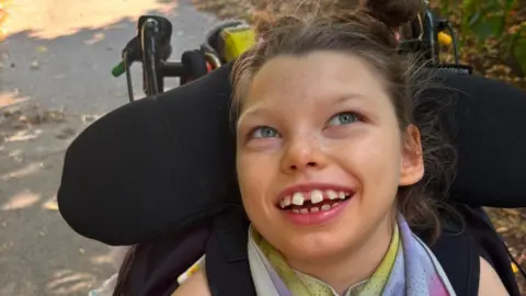 Esmai smiles as she sits in her wheelchair. She has grey eyes and light brown, tied-back hair.