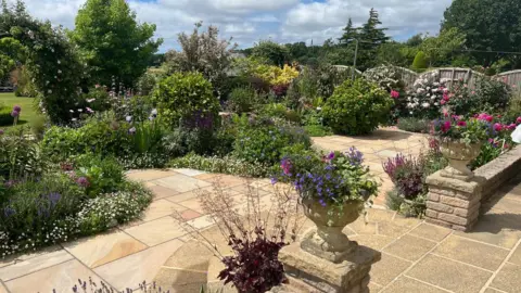 David Hines/Liz Kelly-Hines A large stone paved garden area with lots of colourful greenery and landscaped bushes.