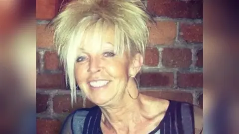 A woman with mid-length blonde hair smiles as she stands in front of a brick wall.