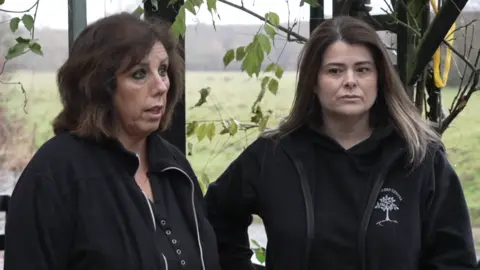 Sue Proctor and Sarah Baxter, both wearing black jackets and tops, stand in their outdoor cafe with leaves around and a river underneath. Fields and hills can be seen on the other side of the river.