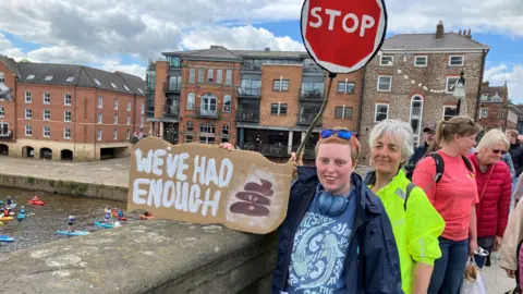BBC/Olivia Richwald A group of people standing on a stone bridge beside a river. In the foreground, one individual holds a handmade cardboard sign with bold white lettering that reads, “WE’VE HAD ENOUGH”, along with a small painted illustration of faeces. Another sign shaped like a red stop sign with the word “STOP” in white capital letters is held up on a stick above the group.
Behind the people on the bridge, a river flows from left to right, where several individuals are paddling in kayaks and on paddleboards. The water appears slightly brown, and the riverbank is lined with old brick buildings featuring large windows and modern balconies.
