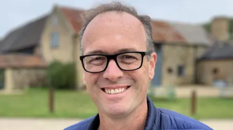 Luke Deal/BBC A head and shoulders photo Hektor Rous. He has short greying hear and black glasses. He is wearing a blue padded jacket and a light green top underneath, and he is stood in front of an old farm building